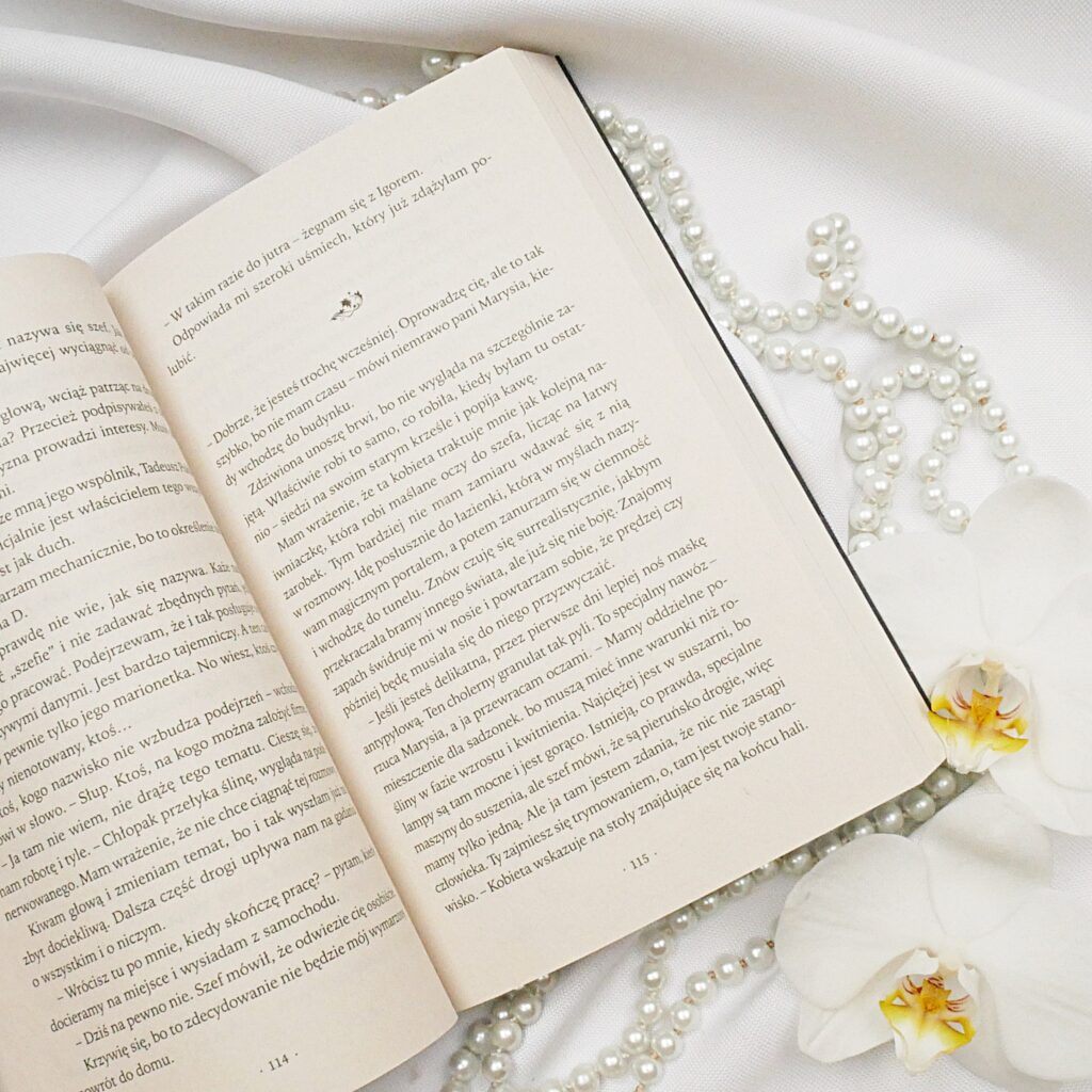 Fragment powieści „Alicja. Po drugiej stronie przyjemności” – Anna Langner Otwarta książka „Alicja. Po drugiej stronie przyjemności” Anny Langner na tle białej tkaniny z perłami i kwiatami orchidei