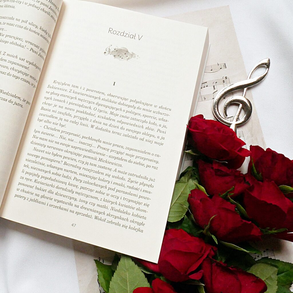 Recenzja książki „Wyśniony koncert” Anny Balenkowskiej Rozdział V książki „Wyśniony koncert” Anny Bałenkowskiej, obok ozdobne nuty, czerwone róże i klucz wiolinowy.