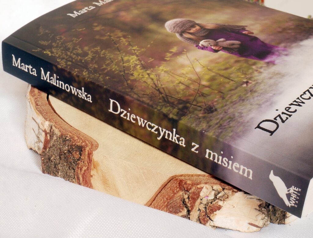 Grzbiet i okładka książki „Dziewczynka z misiem” Marty Malinowskiej Grzbiet książki „Dziewczynka z misiem” Marty Malinowskiej, leżącej na drewnianym plastrze.